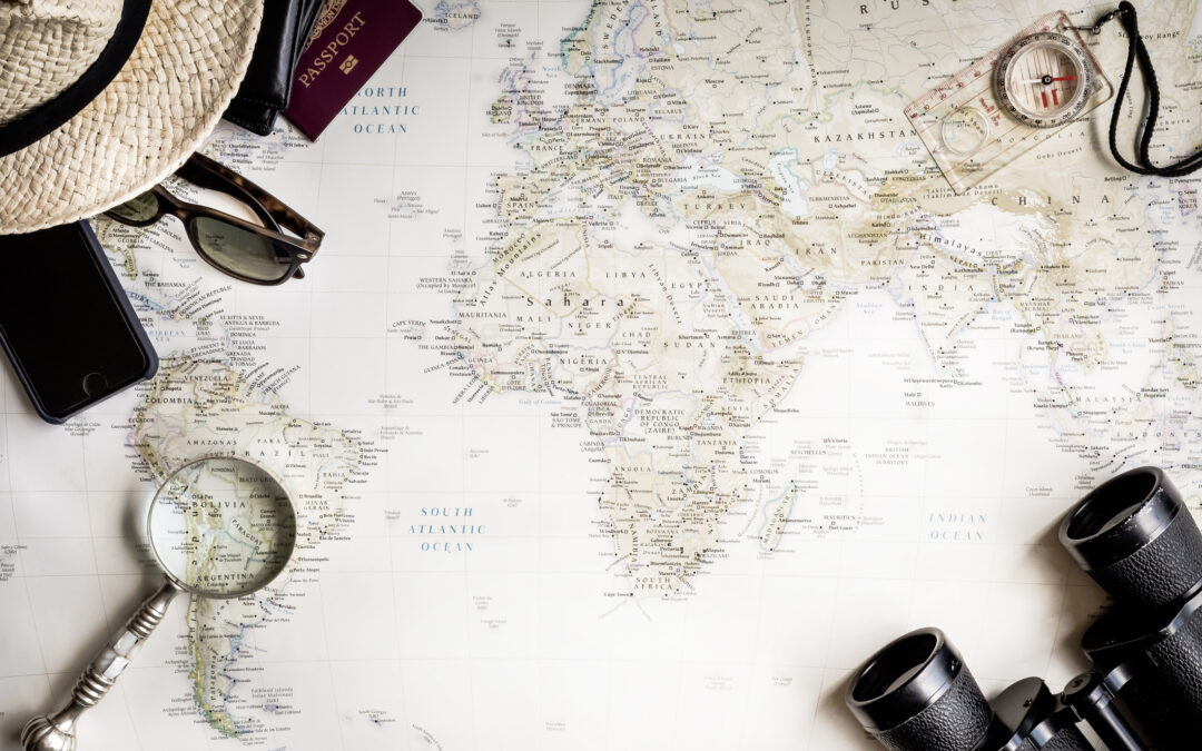 Flat lay, top-view arrangement of travel essentials including a folded map, sunglasses, a camera, a passport, and small accessories neatly placed on a light-colored surface, creating a styled travel-planning scene.