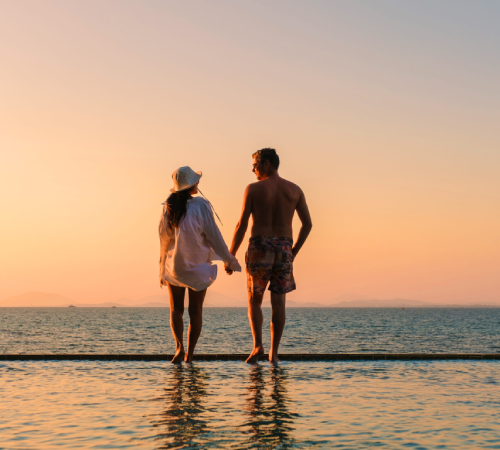 Couple holding hands while walking beside a serene pool at sunset on Koh Munnork Island, Thailand, with golden light reflecting on tranquil waters for a romantic atmosphere.