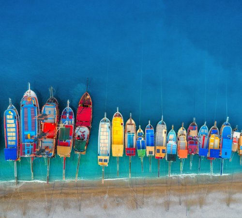 Aerial drone view of colorful boats and yachts floating in clear azure waters along the sandy beach of Oludeniz, Turkey, capturing a vibrant Mediterranean summer seascape.