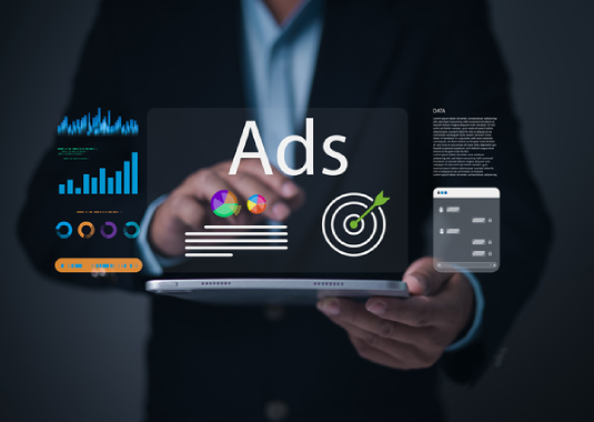 Business professional standing in the background while managing and posting digital ads on a computer, with charts and marketing materials visible, illustrating active online advertising and campaign strategy.