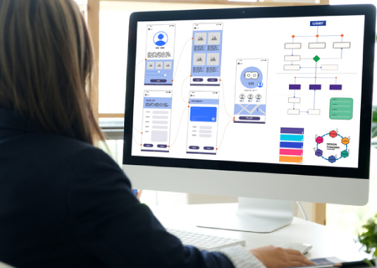 Woman sitting at her desk, focused on website development and design strategies on her computer, with sketches, notes, and a tablet nearby, illustrating creative web planning and digital design work.