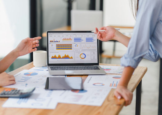 Team gathered around a laptop, discussing analytics and reviewing dashboard data, with charts and graphs visible, illustrating collaborative data analysis and business insights.