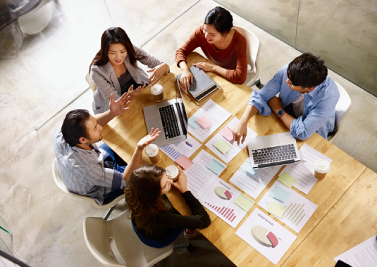 Aerial view of a full-service marketing team gathered around a large table, collaborating on strategies with laptops, notebooks, charts, and coffee cups, showing teamwork and a dynamic brainstorming session.